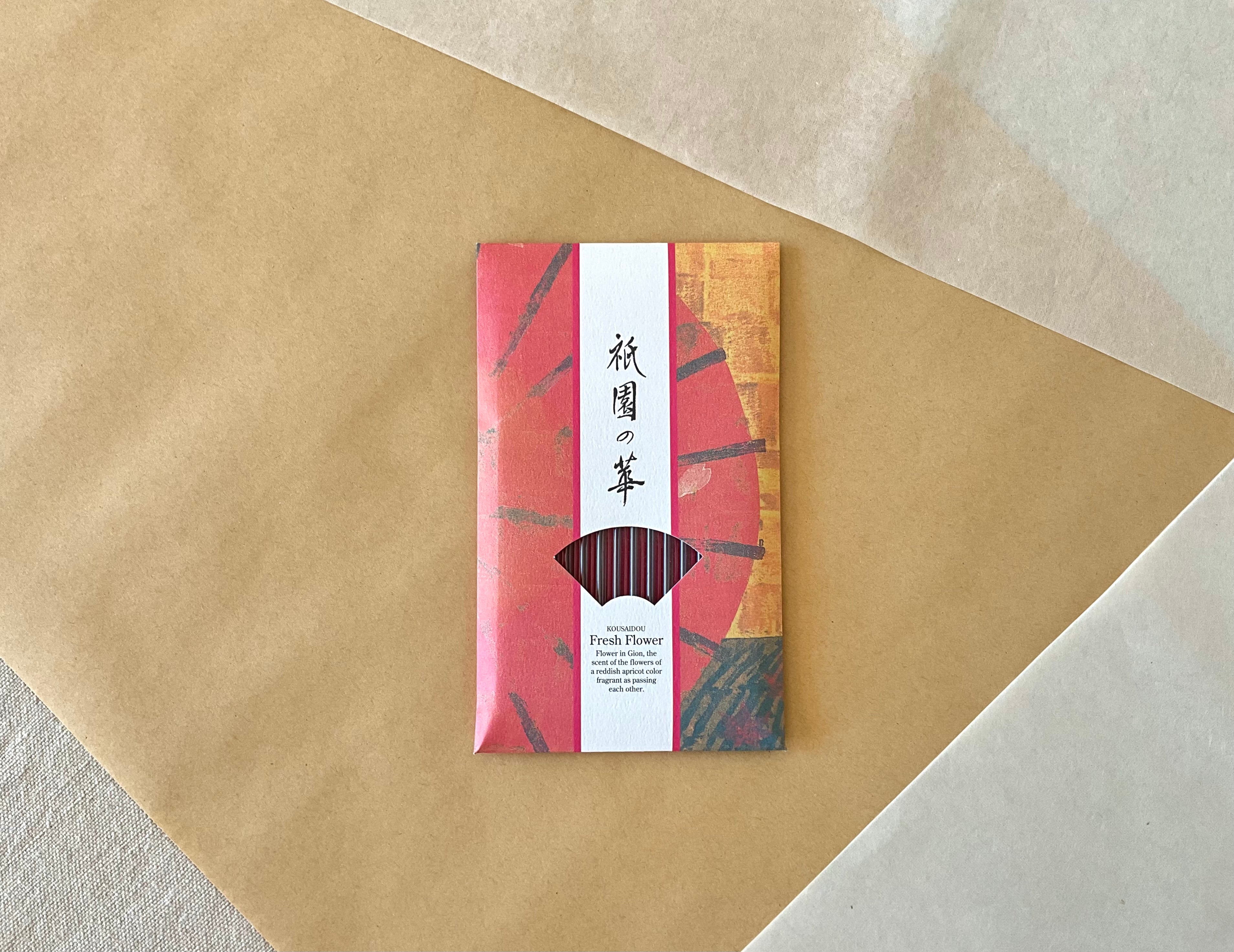 Kousaidou Gion Fresh Flower incense pack centered on diagonal kraft paper background, full red and gold illustrated sleeve with Gion district design, fan window showing dark reddish incense sticks, Kousaidou and Fresh Flower text in English