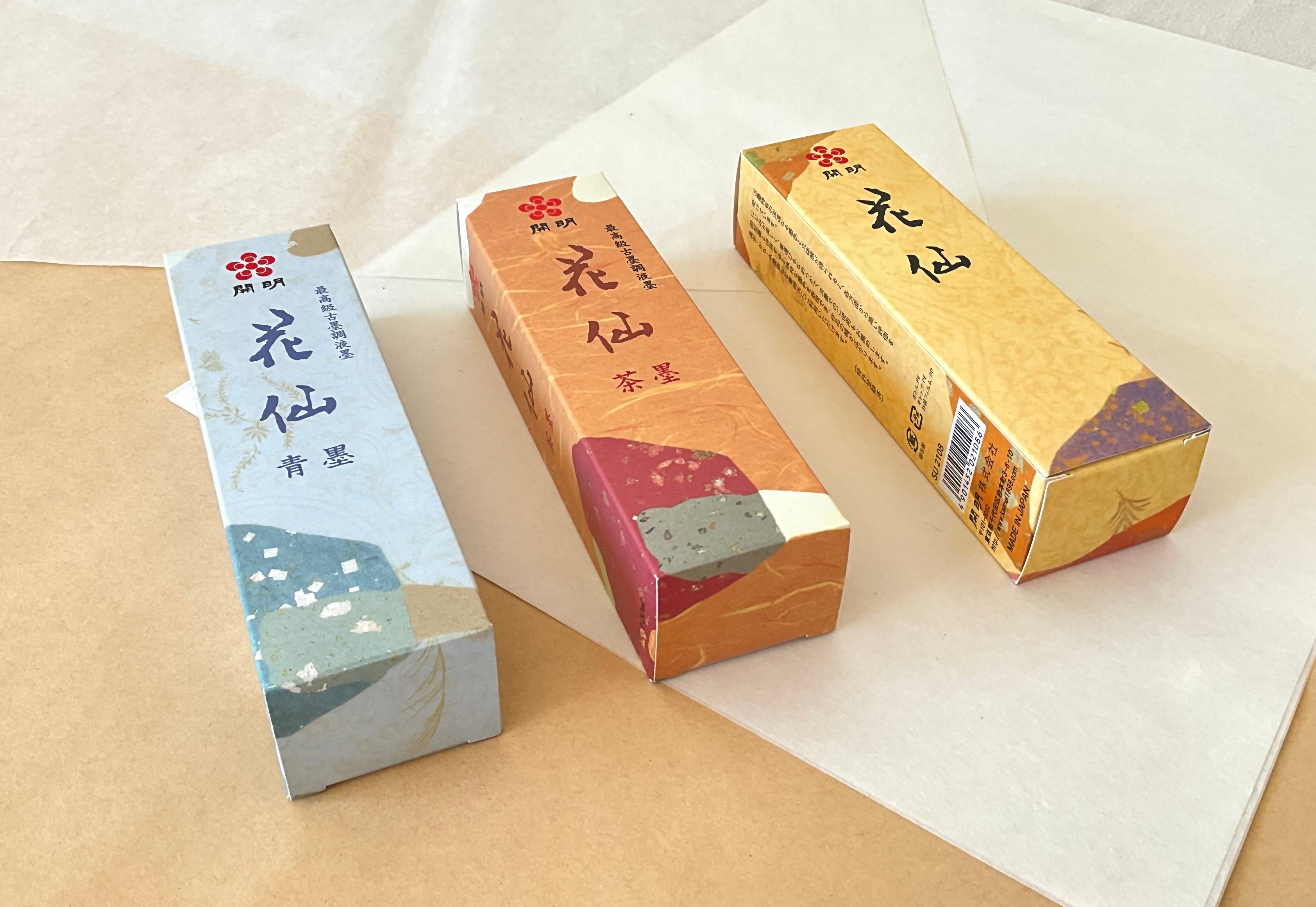 Three Kaimei Kasen premium liquid ink boxes side by side, Seiboku blue-black, Chaboku brown-black, and standard black,on washi and kraft paper background