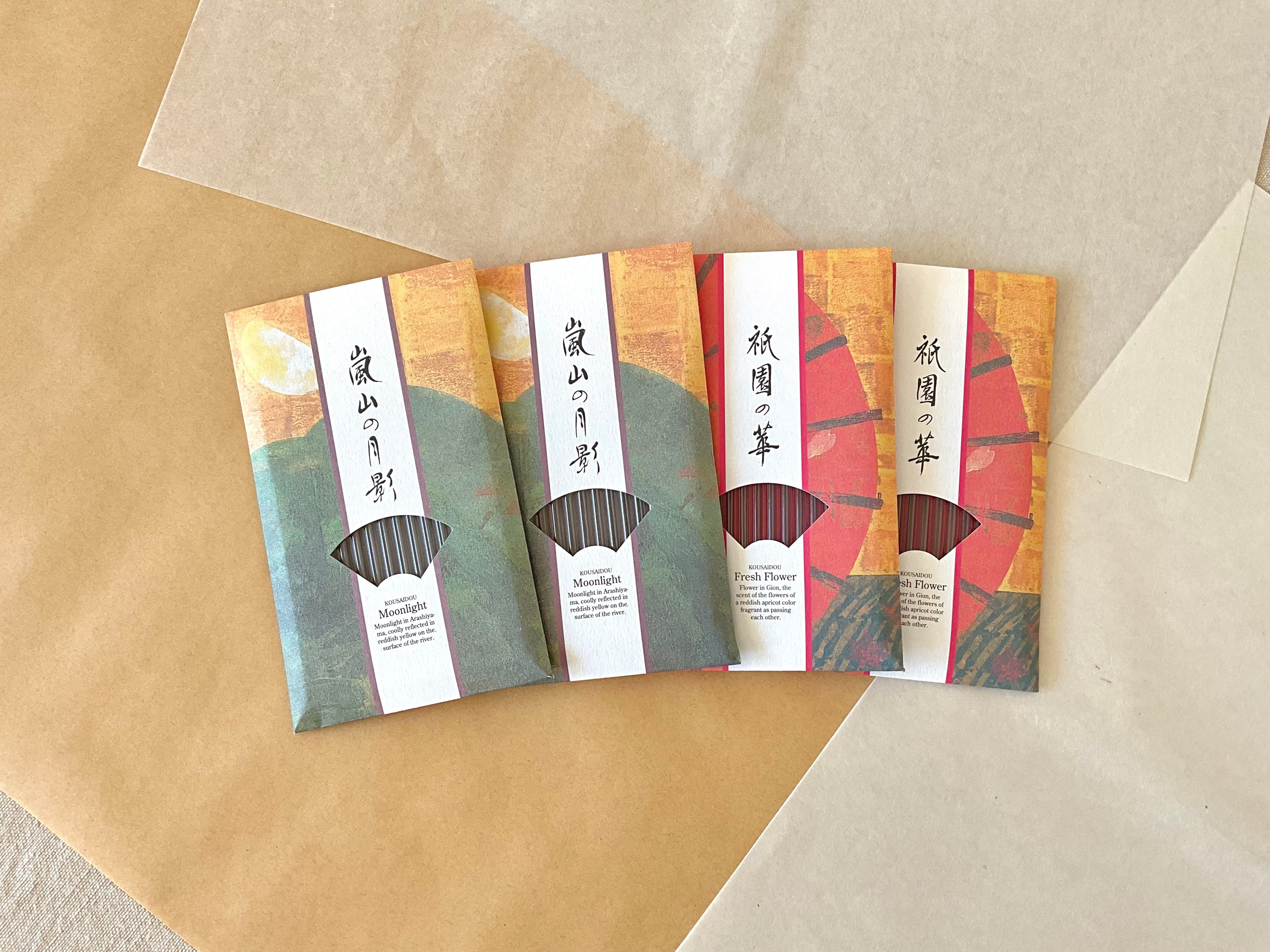 Four Kousaidou Kyoto incense packs fanned on kraft paper showing two Gion Fresh Flower packs in red and gold with abstract Gion illustration, and two Arashiyama Moonlight packs in green and gold, calligraphy titles and fan window showing incense sticks