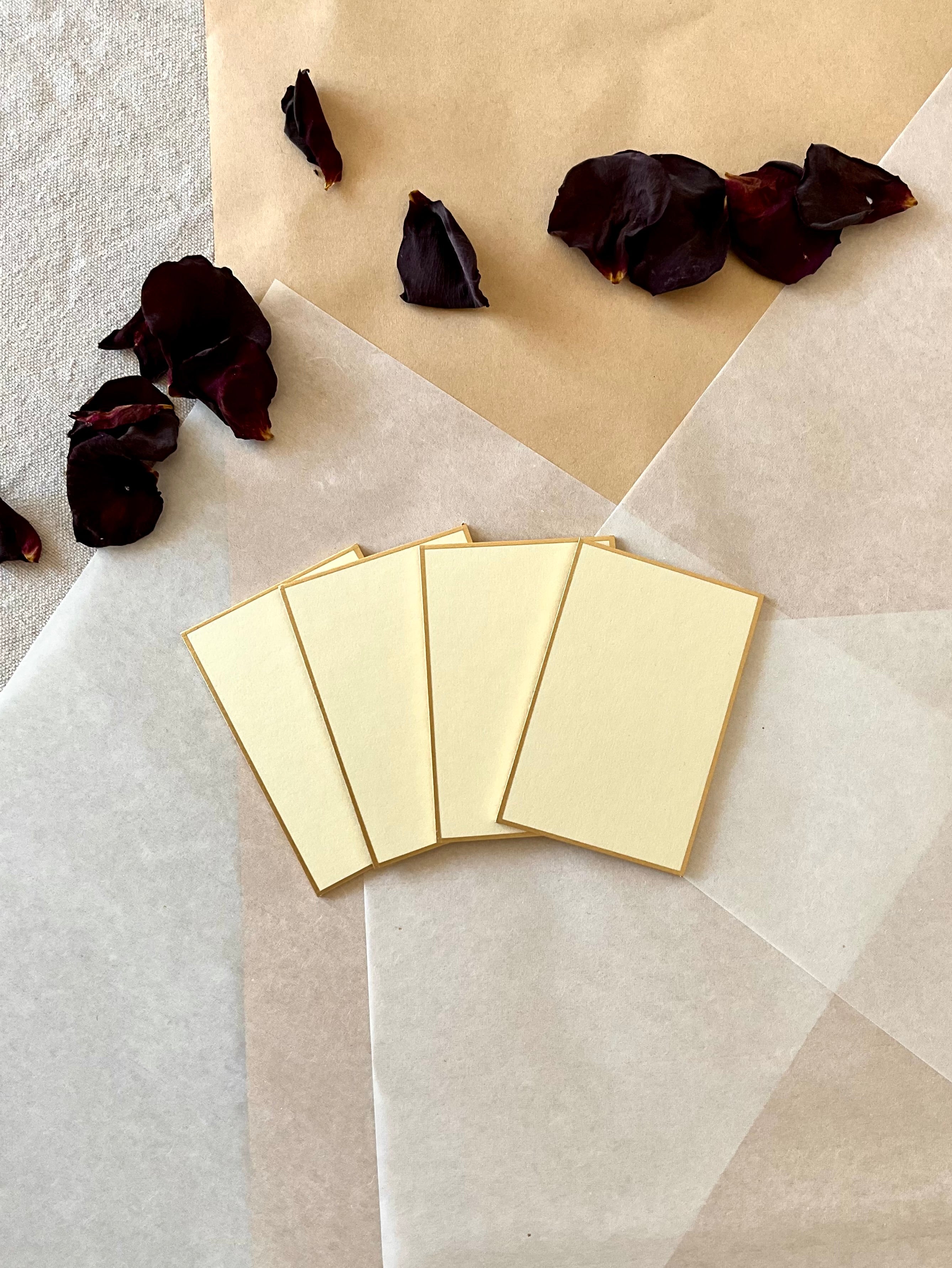 Four small rectangle cream Kyukyodo shikishi boards fanned out, centered on layered washi and kraft paper with dried dark rose petals at upper left, showing the clean rectangle form and gold-painted edge
