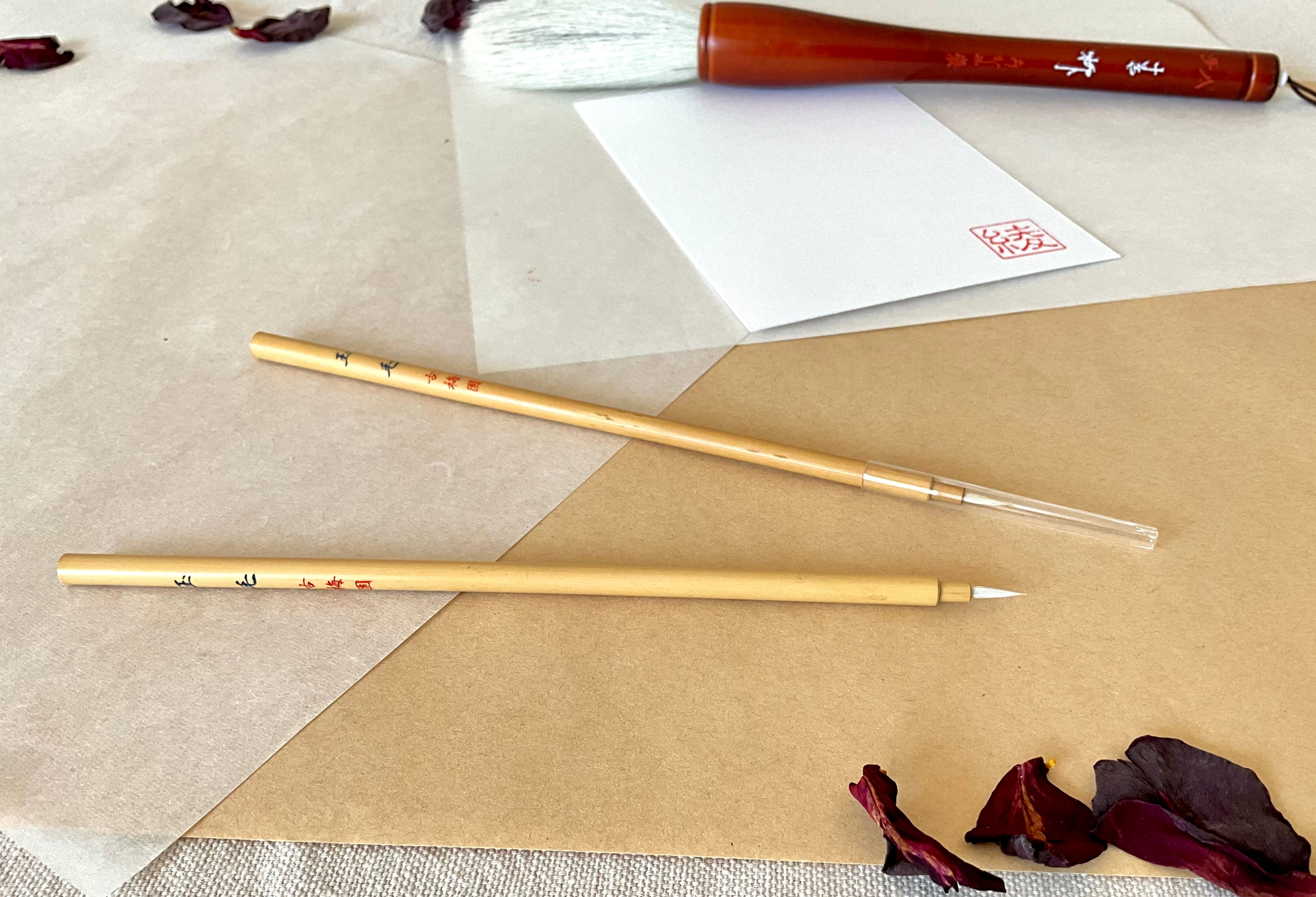 Two Kobaien fine detail calligraphy brushes with sharp pointed tip and bamboo handle, one covered with a lid and one is open, resting on layered washi paper with dried rose petals and a red hanko stamp card