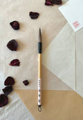 Kobaien large calligraphy brush with firm tapered bristle, black lacquered ferrule and pale bamboo handle with printed paper label and hanging loop, resting on layered washi paper with dried rose petals and a red hanko stamp card