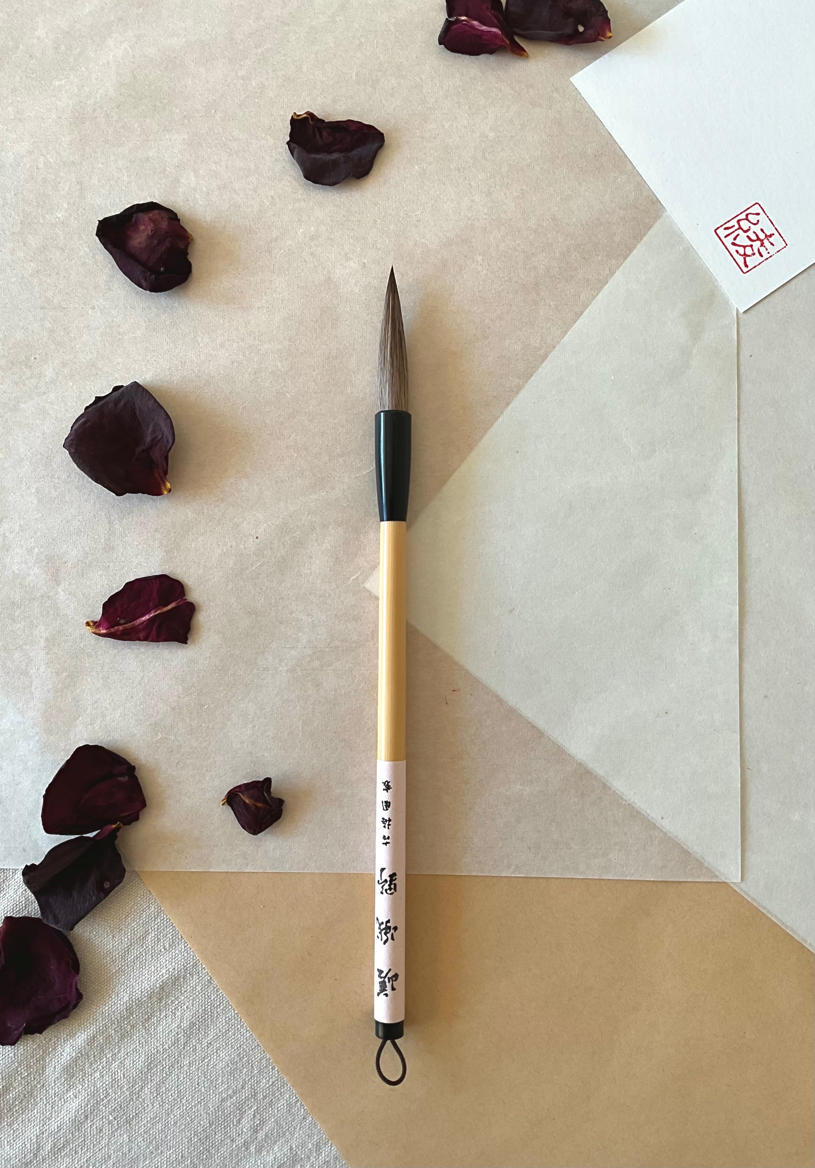 Kobaien large calligraphy brush with firm tapered bristle, black lacquered ferrule and pale bamboo handle with printed paper label and hanging loop, resting on layered washi paper with dried rose petals and a red hanko stamp card
