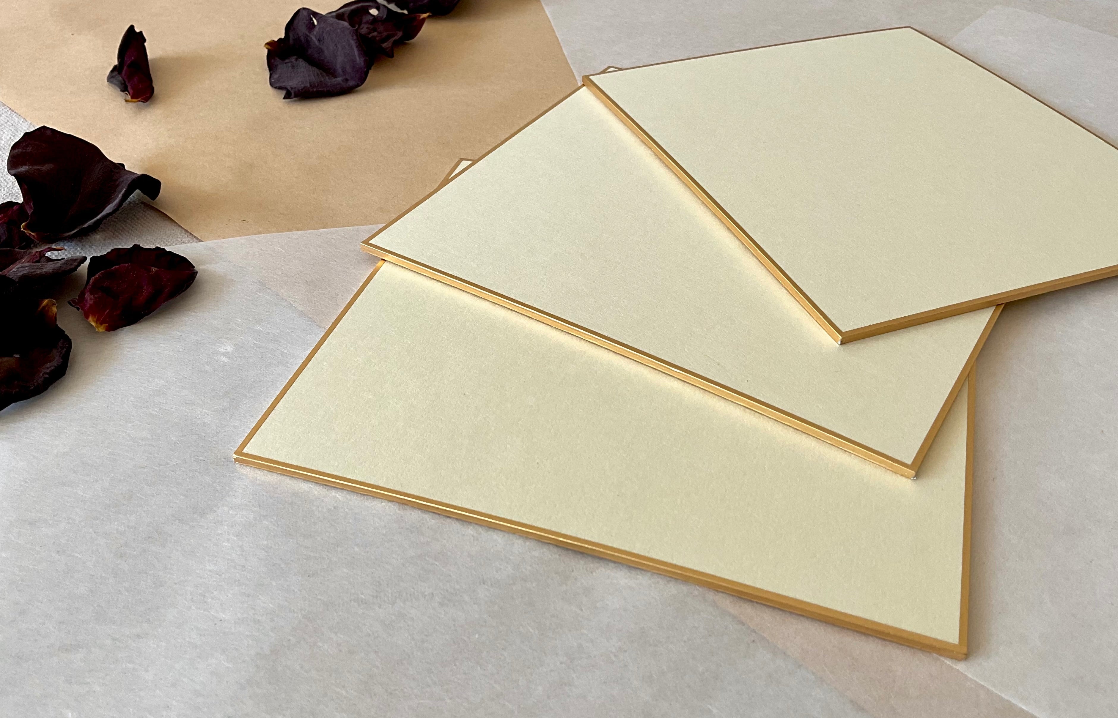 Three large cream Kyukyodo shikishi boards fanned out at an angle on layered washi and kraft paper with dried dark rose petals to the left, showing the ivory surface and fine gold border