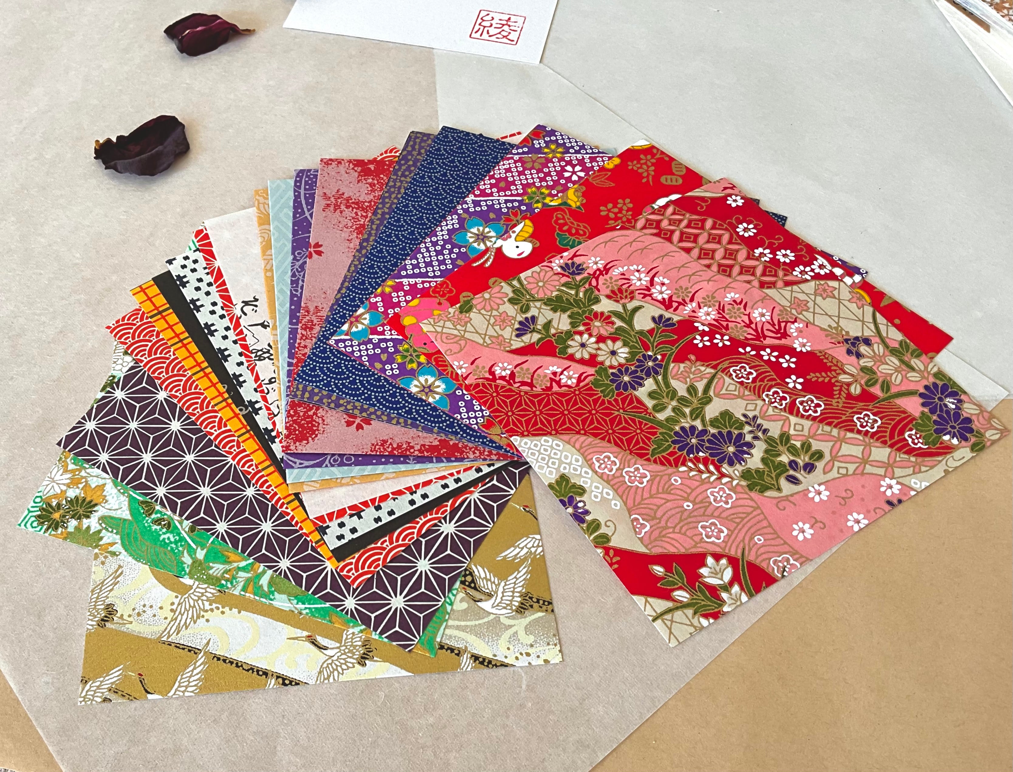 Open pack of Morita Washi chiyogami origami paper fanned out showing all 20 mixed design sheets including crane motifs, navy shibori dots, geometric asanoha star pattern in aubergine, red wave florals and kimono patchwork patterns, on washi paper background