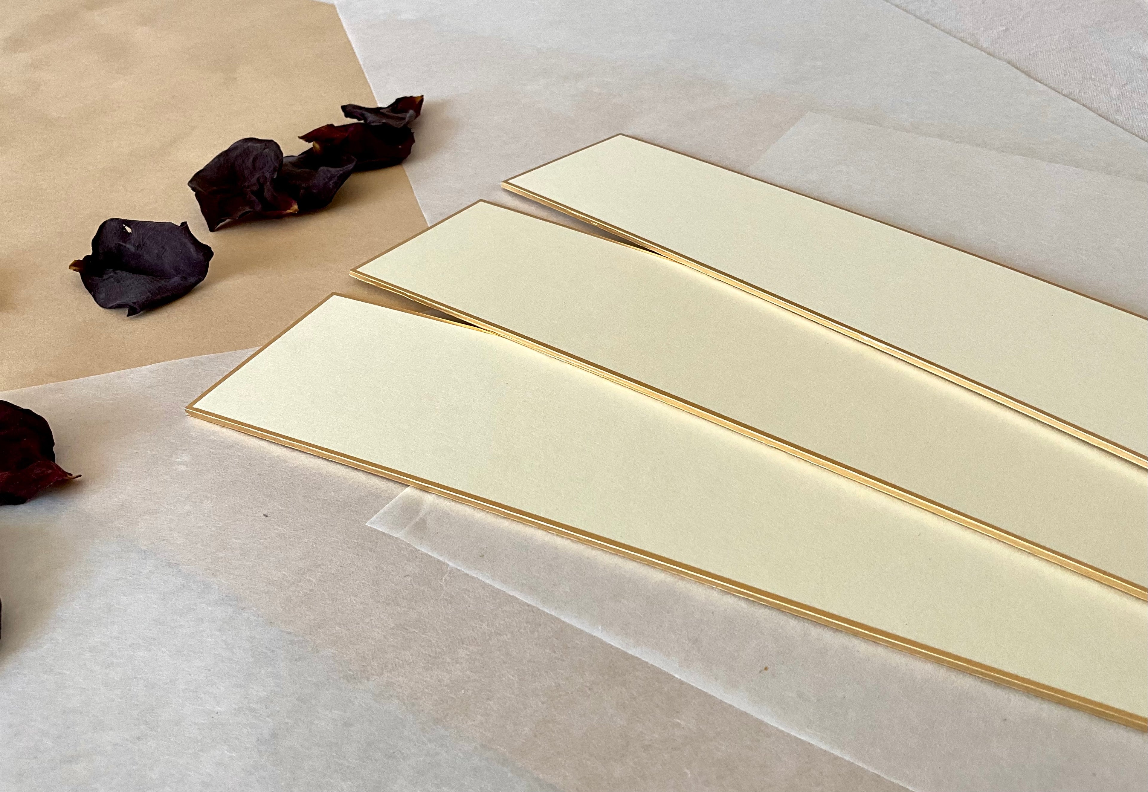 Three cream Japanese tanzaku strips with gold edges fanned out at an angle on washi paper, dried rose petals scattered to the left, showing the smooth ivory surface and fine gold border