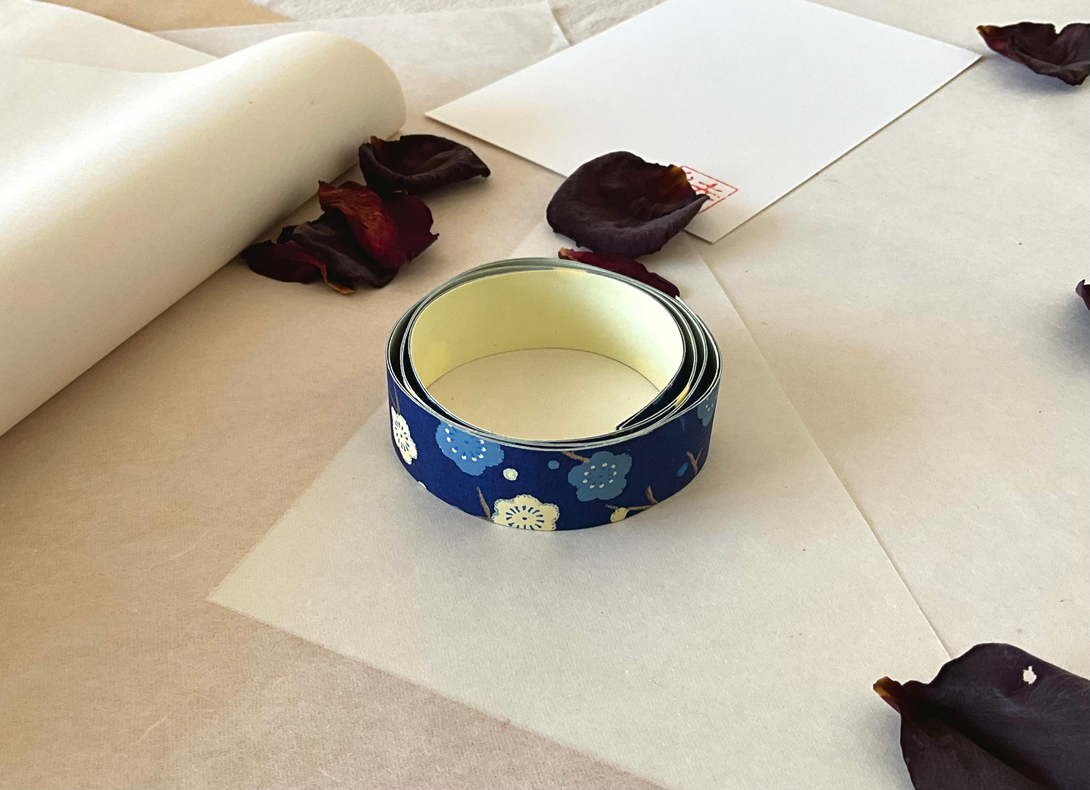 Shogado yuzen washi tape unrolled on washi paper showing Navy with cream and pale blue plum blossom branches along its length, adhesive backing visible, dried rose petals nearby