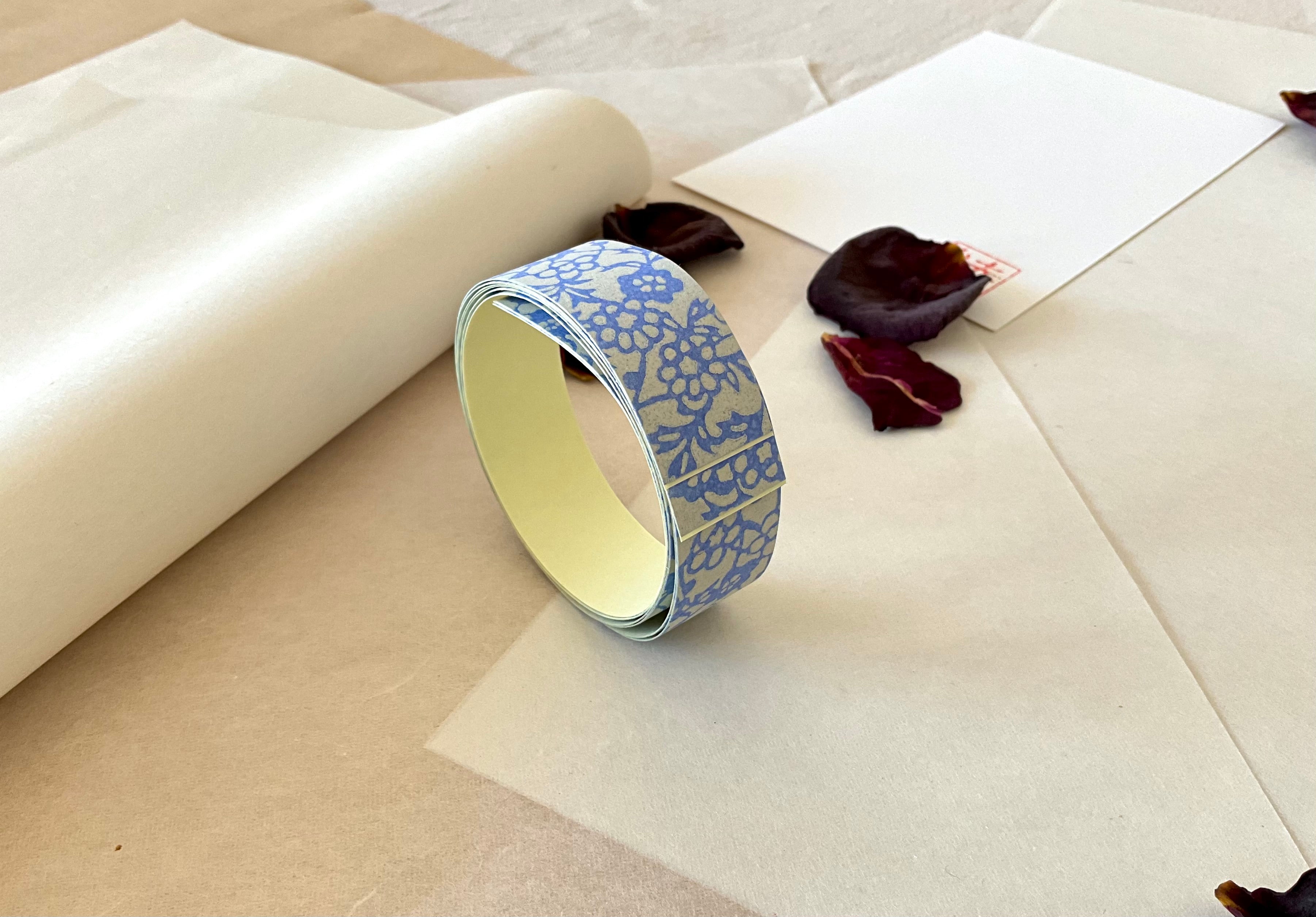 Roll of Shogado hand dyed yuzen washi tape standing upright on layered washi and kraft paper, Grey-blue with blue floral block print pattern, cream adhesive backing visible, dried rose petals and hanko stamp card in background