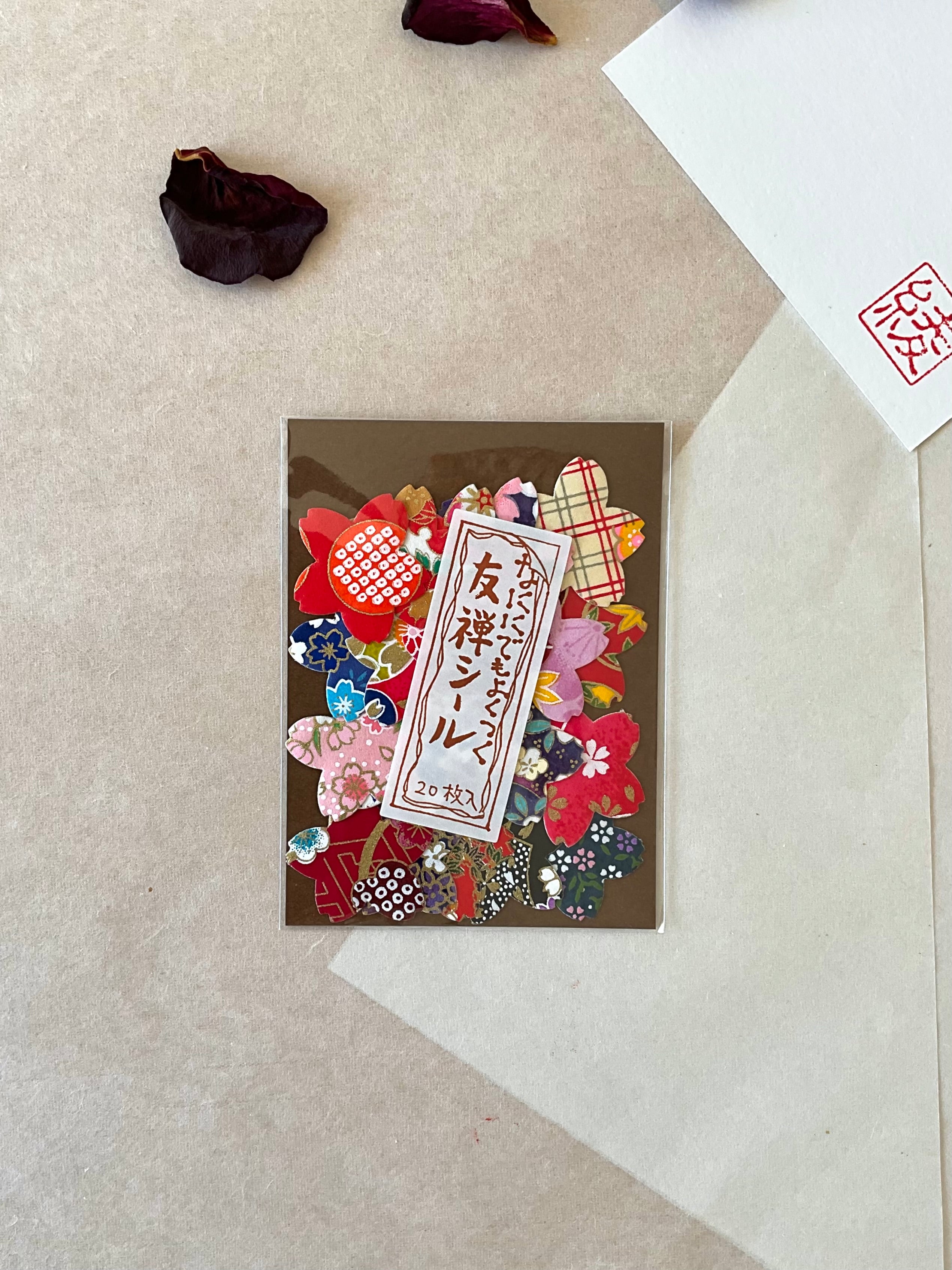 Single Morita Washi yuzen sticker pack from directly above on neutral washi background, showing 20 mixed flower shaped stickers in traditional Japanese patterns covering the full brown card backing, with hanko stamp card and dried rose petal