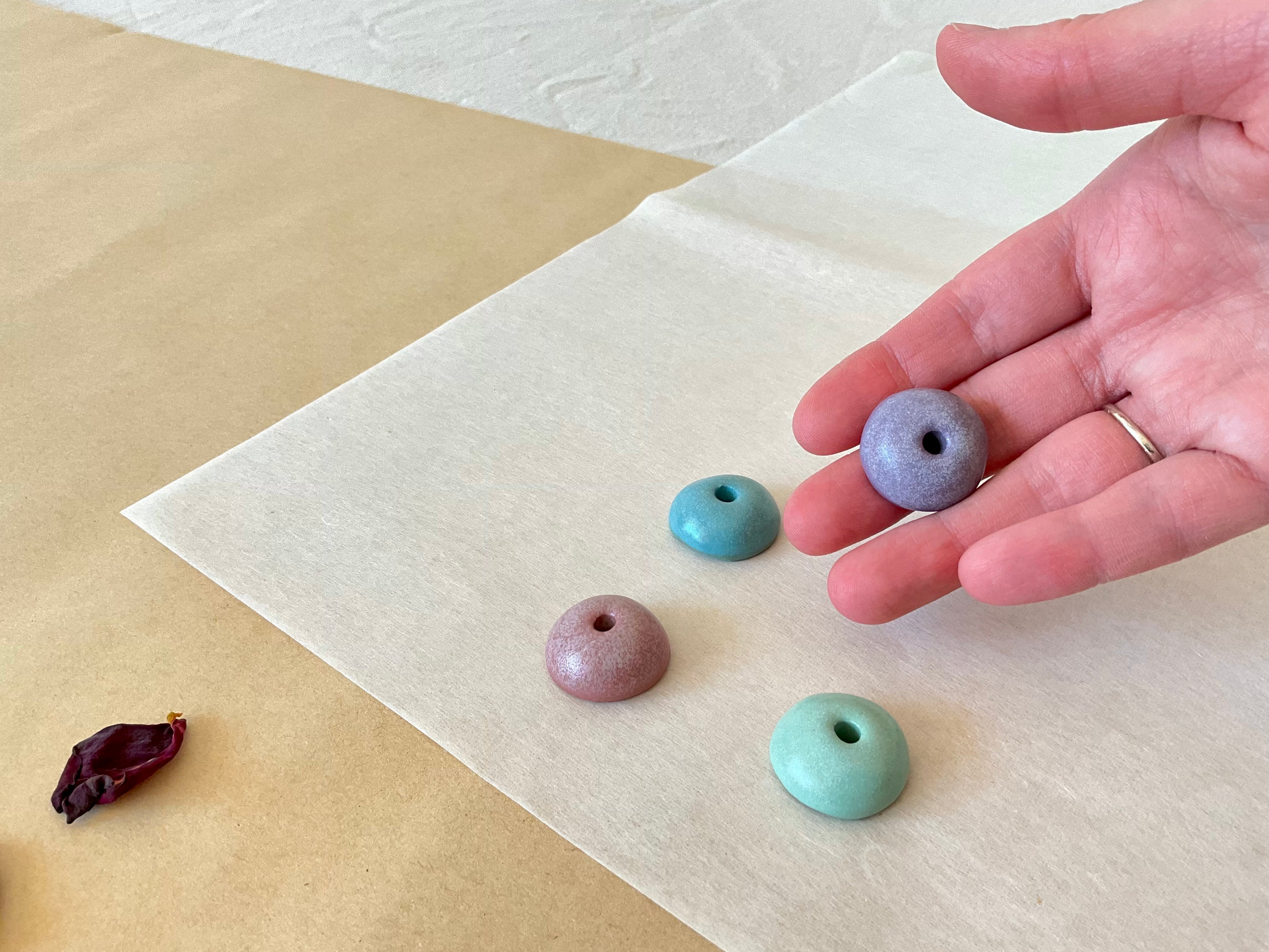 Three round ceramic incense holders resting on washi paper in teal, dusty rose and sage green, with a lavender holder held in an open hand showing the glaze texture and central hole, dried rose petal nearby