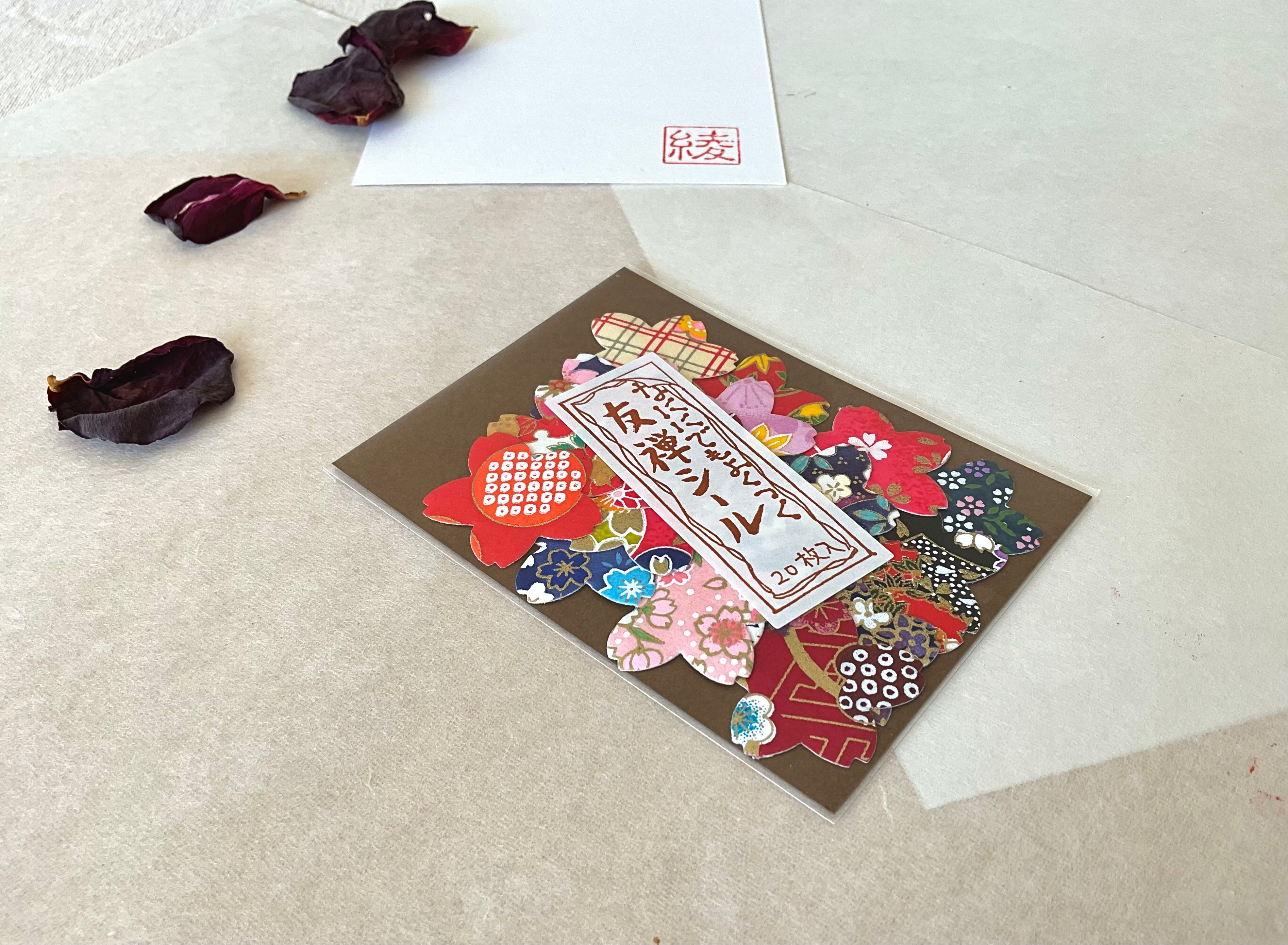 Morita Washi yuzen washi sticker pack tilted on washi paper, showing full color range including red dotted floral, dark navy, pink plaid, gold and purple chiyogami flower shaped stickers, with dried rose petals