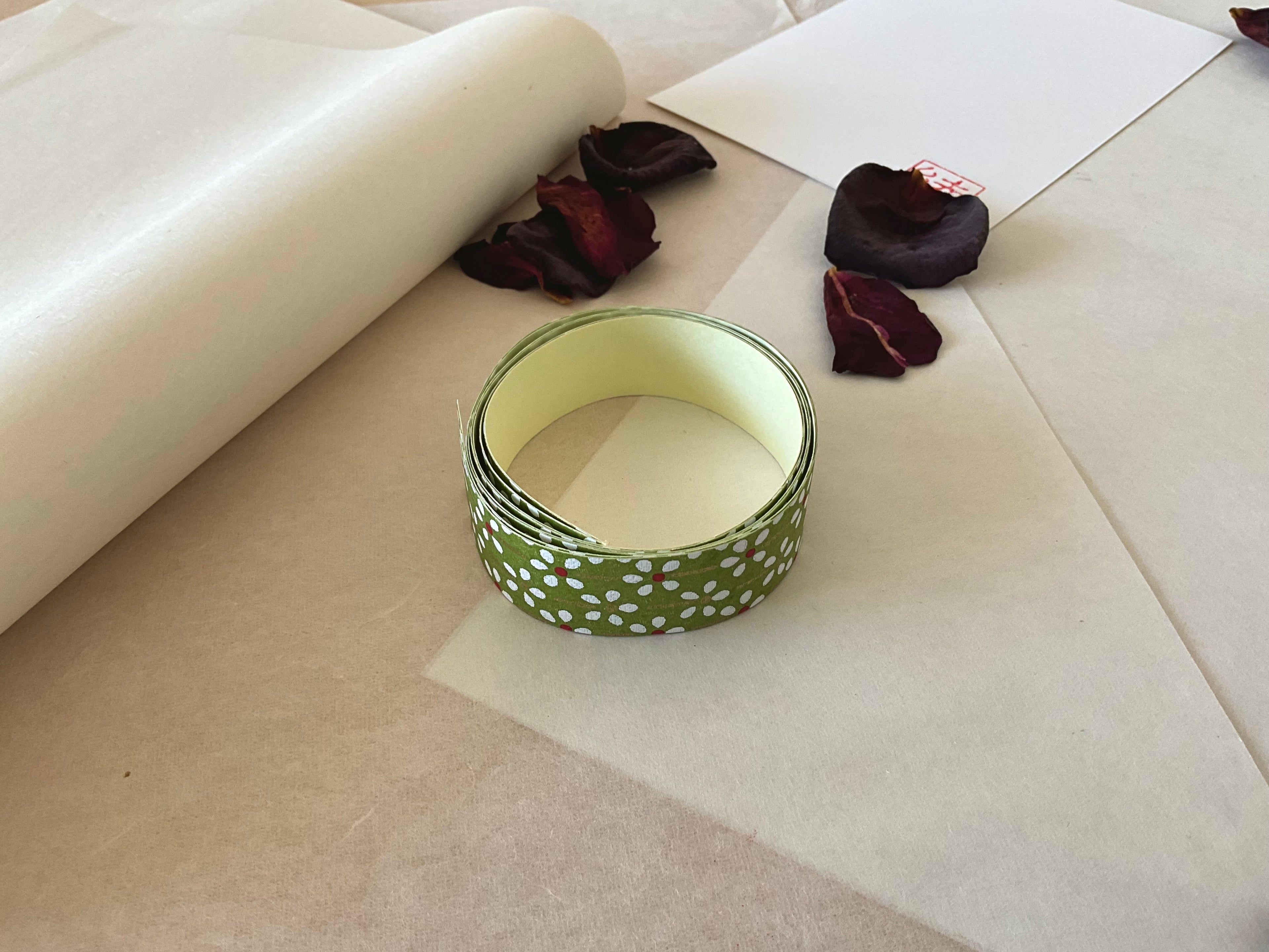 Shogado yuzen washi tape unrolled on washi paper showing Olive green with white daisy and red center floral print along its length, adhesive backing visible, dried rose petals nearby