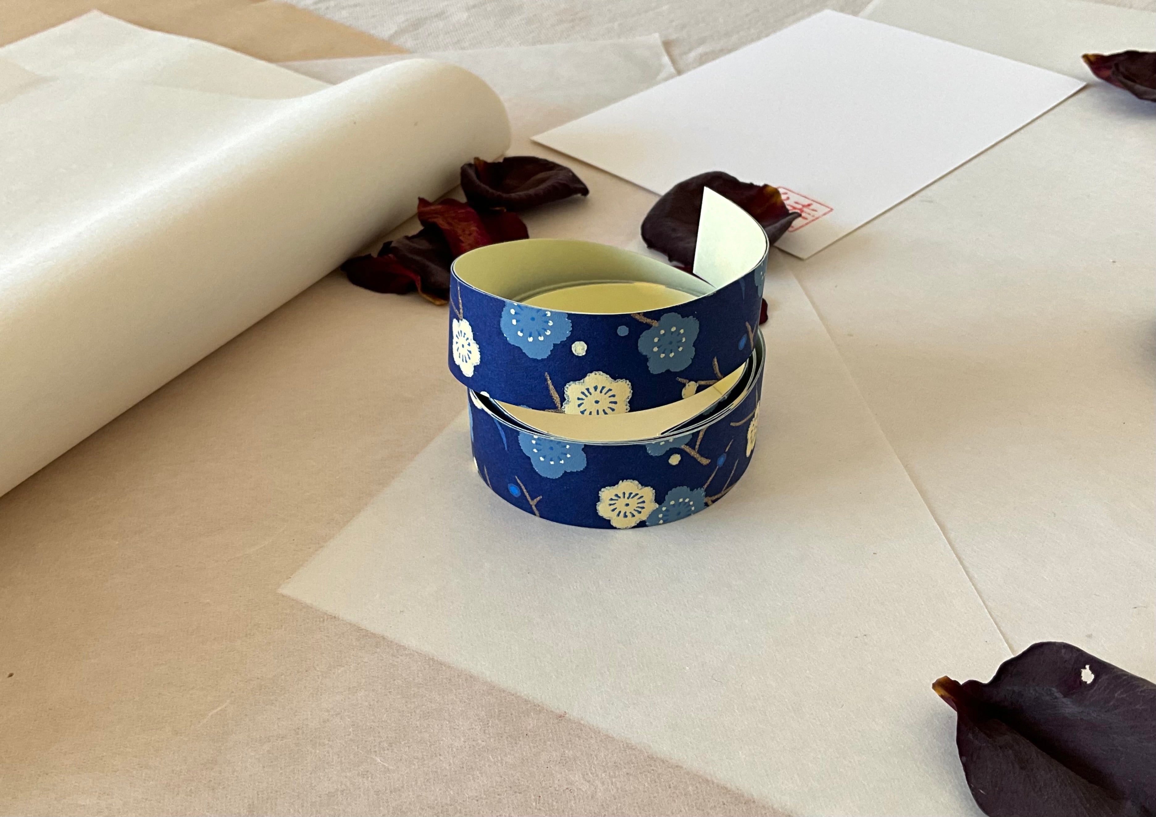 Both sheets of Shogado hand dyed yuzen washi tape laid on top of each other on washi paper, showing Navy with cream and pale blue plum blossom branches repeated across both strips, dried rose petals nearby