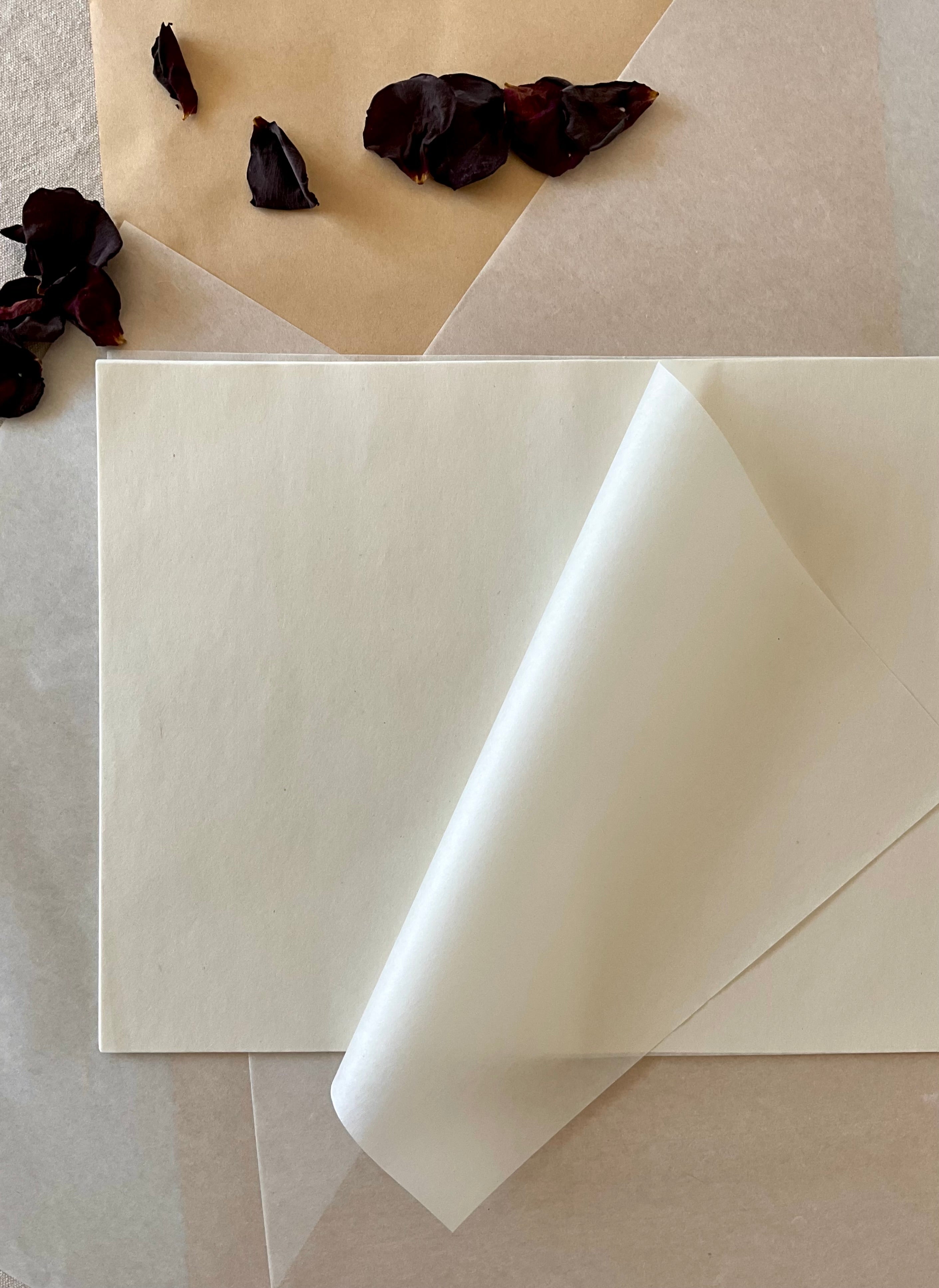 A large sheet of Japanese hanshi paper with one corner curled back, resting on layered washi paper with dried dark rose petals at upper left, showing the soft cream tone and delicate texture of the surface