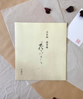 Kyokuodo shikishi calligraphy paper sleeve with Japanese ink brushwork title, resting on layered kraft and washi paper with dried rose petals