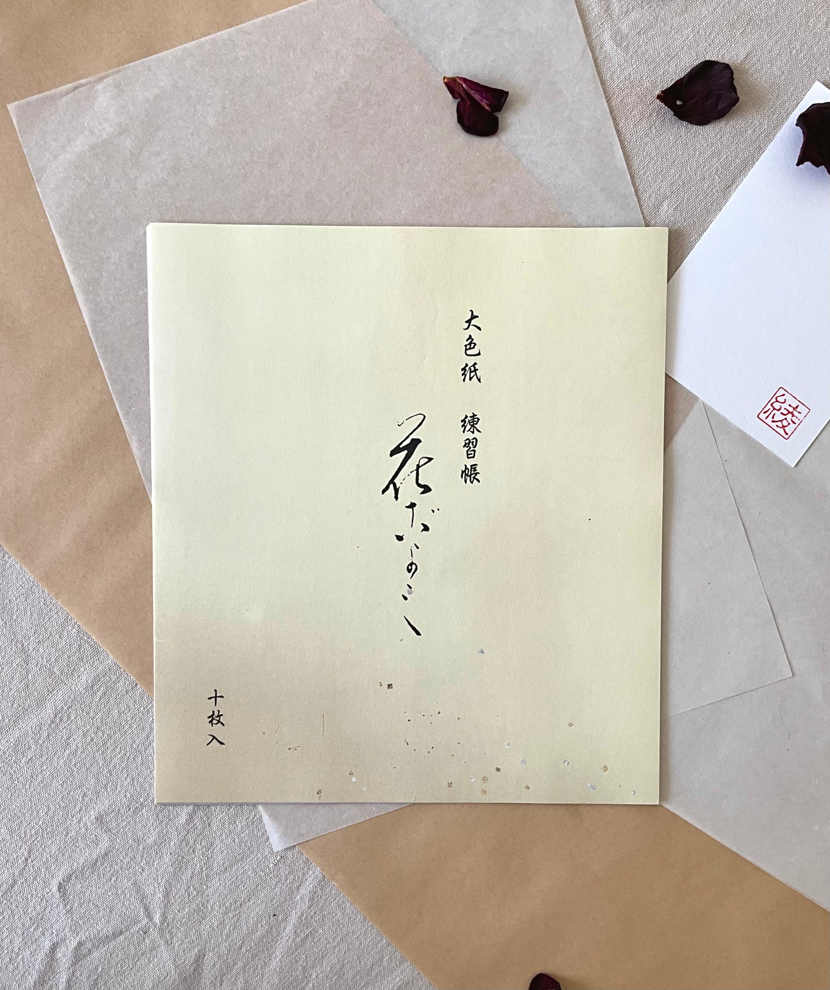 Kyokuodo shikishi calligraphy paper sleeve with Japanese ink brushwork title, resting on layered kraft and washi paper with dried rose petals