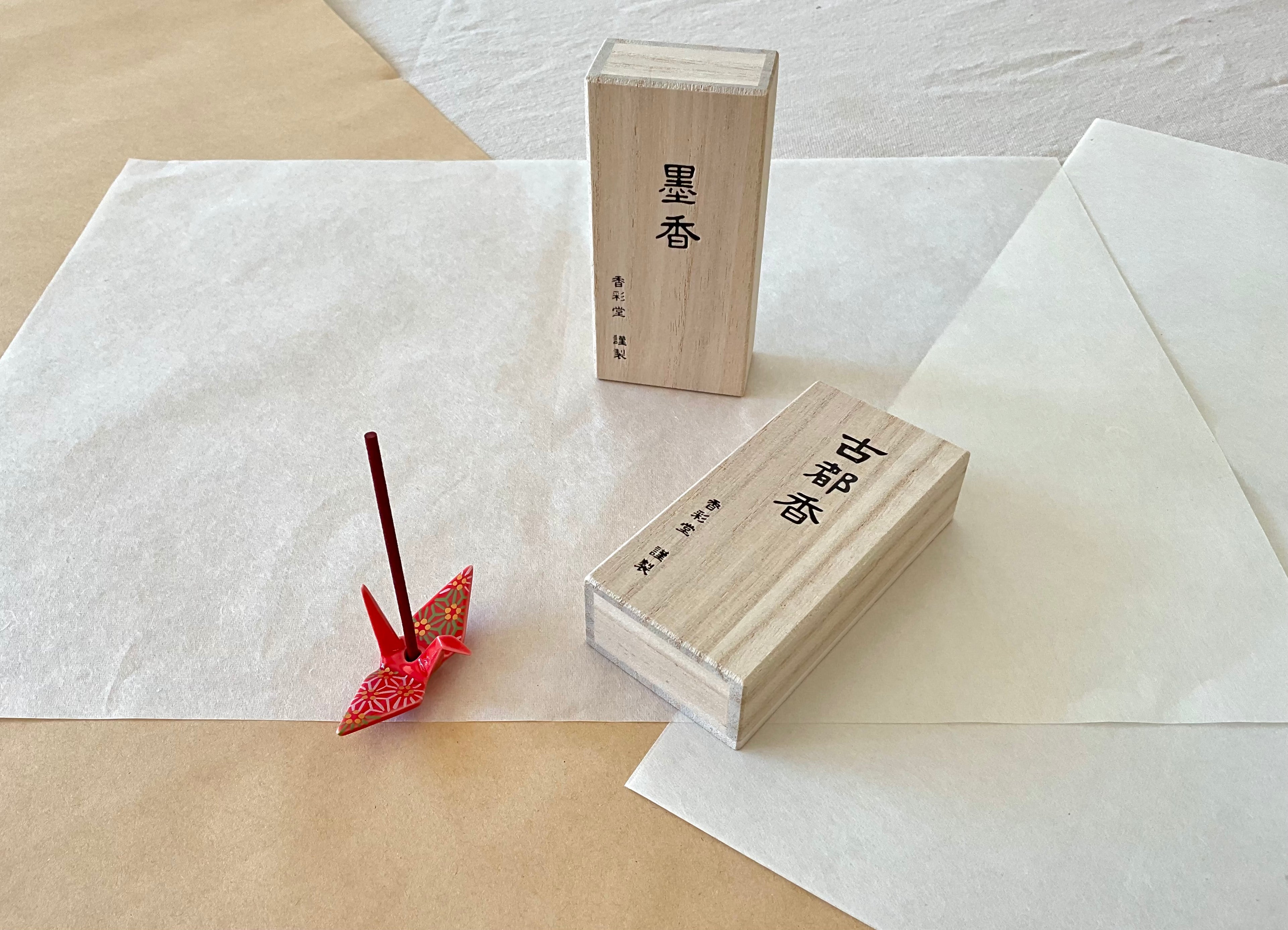 Ceramic origami crane incense holder with red chiyogami pattern glaze standing on washi paper, holding Japanese incense stick, next to a paulownia wood incense box with Japanese kanji