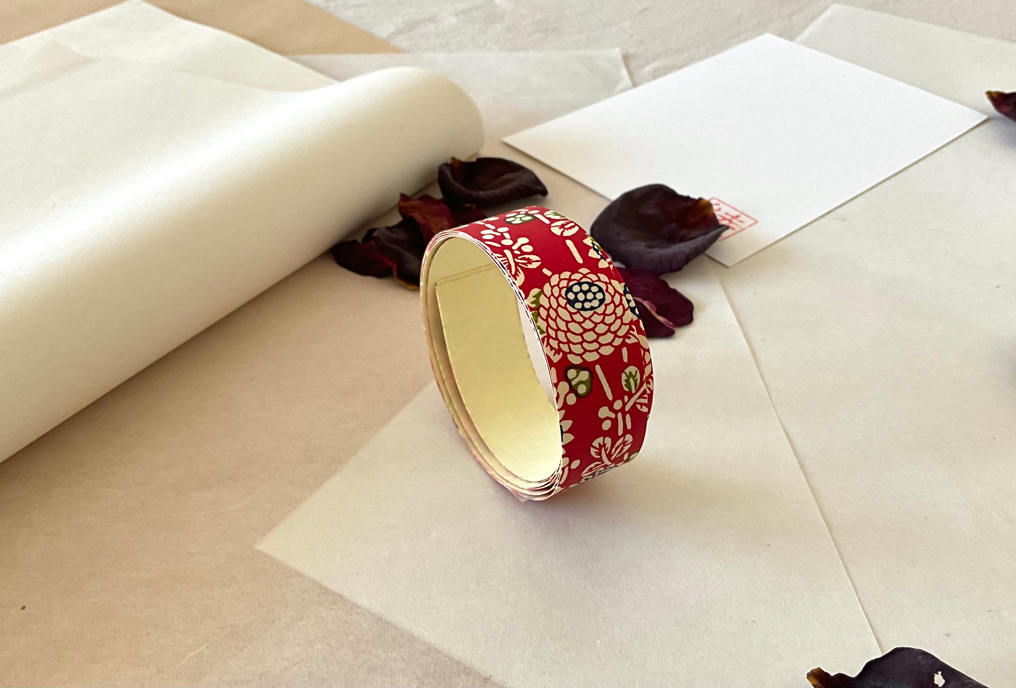 Roll of Shogado hand dyed yuzen washi tape standing upright on layered washi and kraft paper, Crimson red with cream chrysanthemum and floral motifs, navy dotted circle centers, green accents, cream adhesive backing visible, dried rose petals and hanko stamp card in background