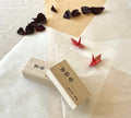 Two boxes one over the other with two red origami cranes on layered washi and kraft paper and washi paper, scattered dried rose petals