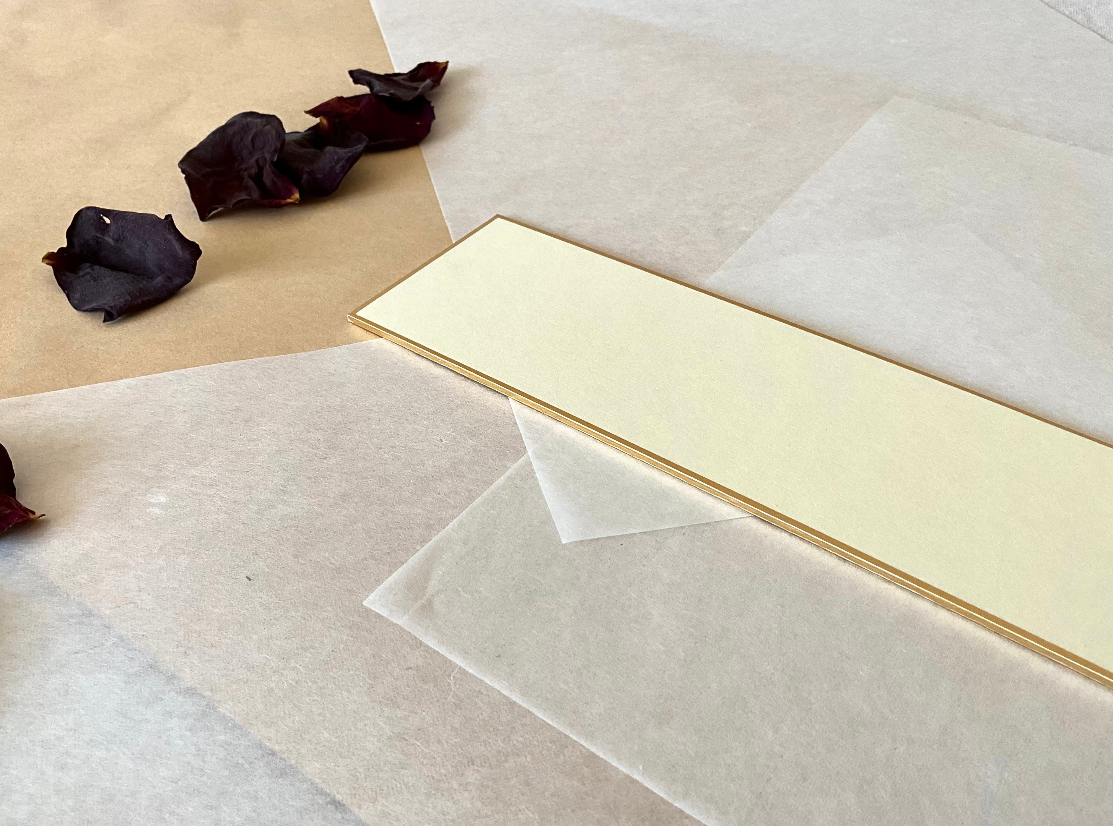 One Japanese tanzaku strips with gold edge, in cream, resting at an angle on layered washi and kraft paper with dried rose petals nearby