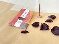 Kousaidou Gion Fresh Flower incense pack leaning on layered kraft and washi paper, bold red and gold illustrated sleeve with Gion calligraphy title, lit dark incense stick in small round pink stone holder, surrounded by scattered dried rose petals