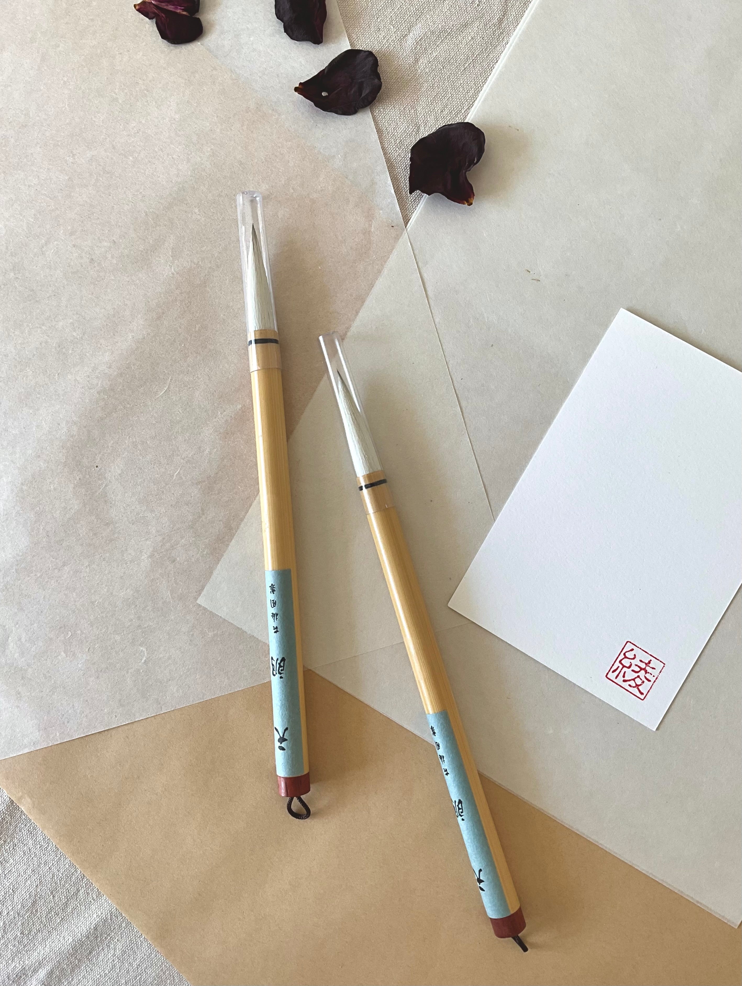 Two Kobaien large calligraphy brushes with pale soft bristle and bamboo handle wrapped in light blue paper, hanging loop at the base, resting on layered washi paper with dried rose petals and a red hanko stamp card