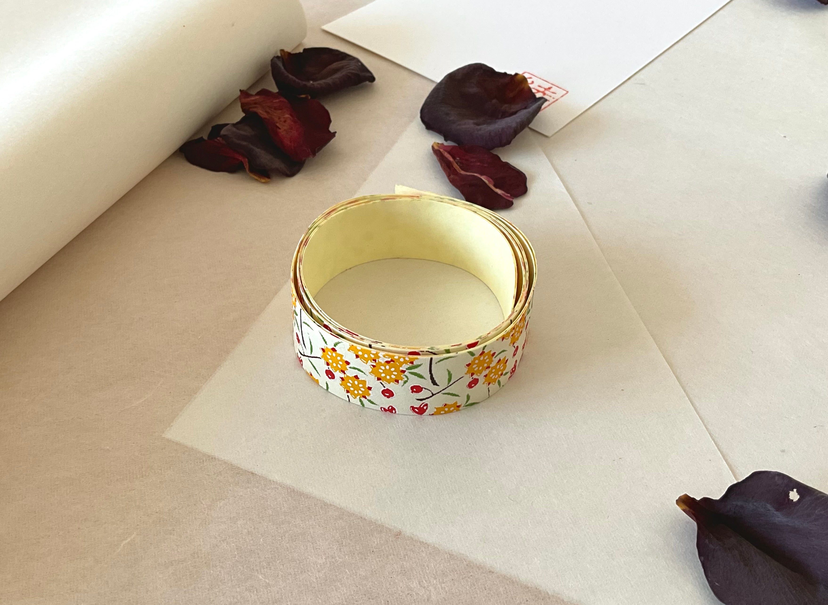 Shogado yuzen washi tape unrolled on washi paper showing Cream with yellow and red floral sprigs along its length, adhesive backing visible, dried rose petals nearby