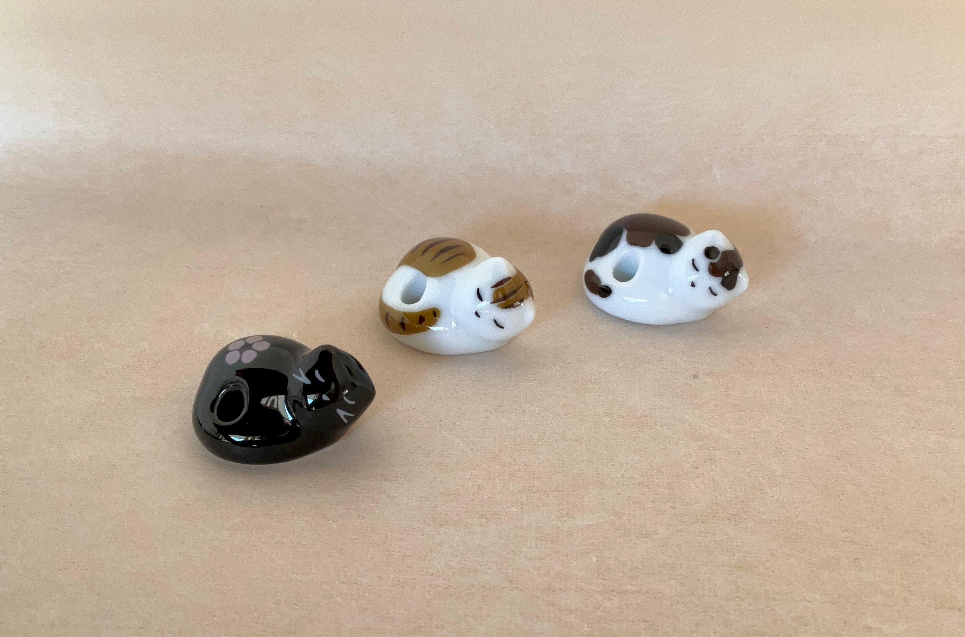 Three ceramic sleeping cat incense holders in a row, one white with brown tabby stripes, one white with black and brown spots, and one black with a floral print, each with a hole along the back for an incense stick