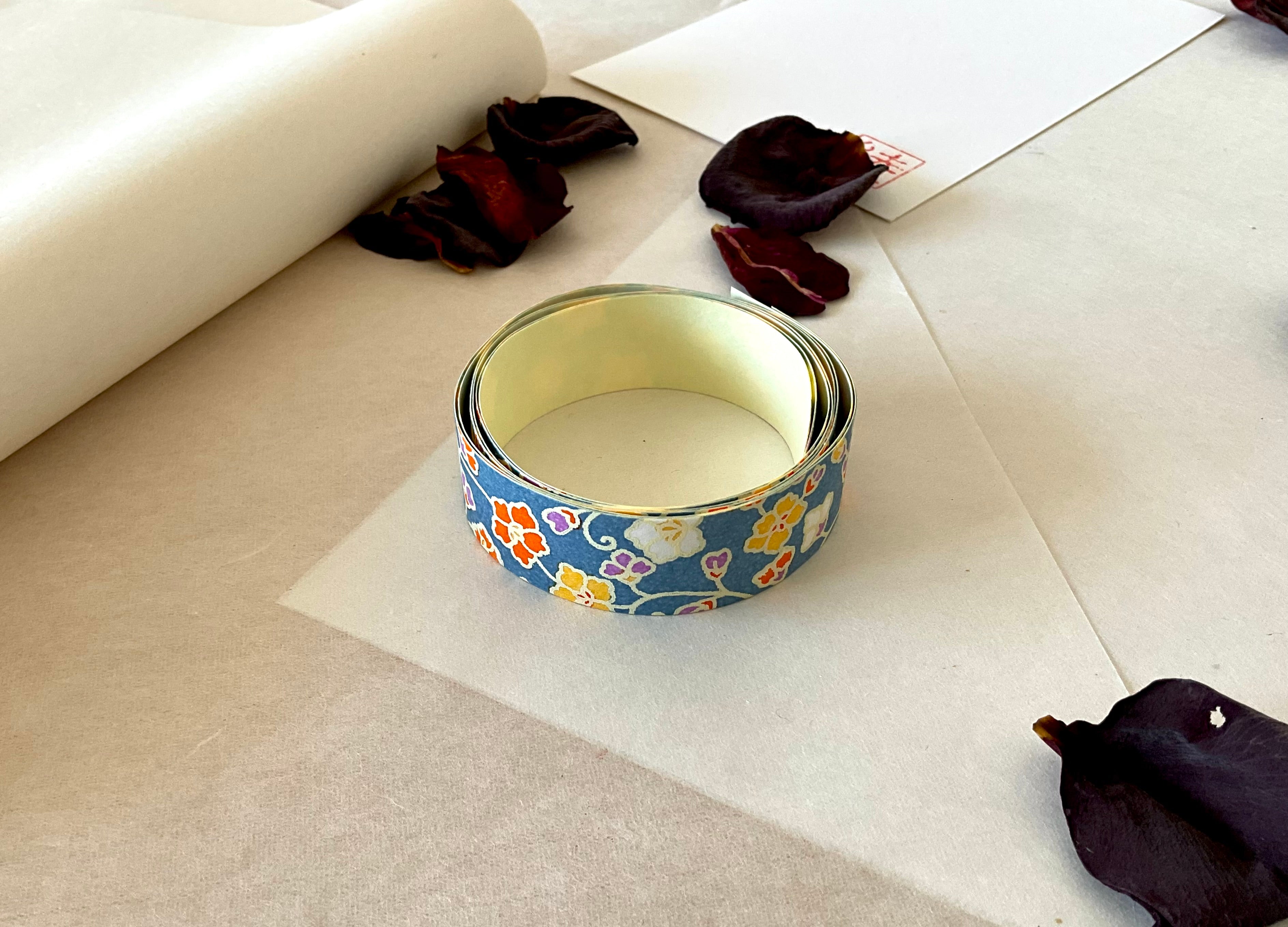 Shogado yuzen washi tape unrolled on washi paper showing Steel blue with yellow, orange, purple small florals along its length, adhesive backing visible, dried rose petals nearby
