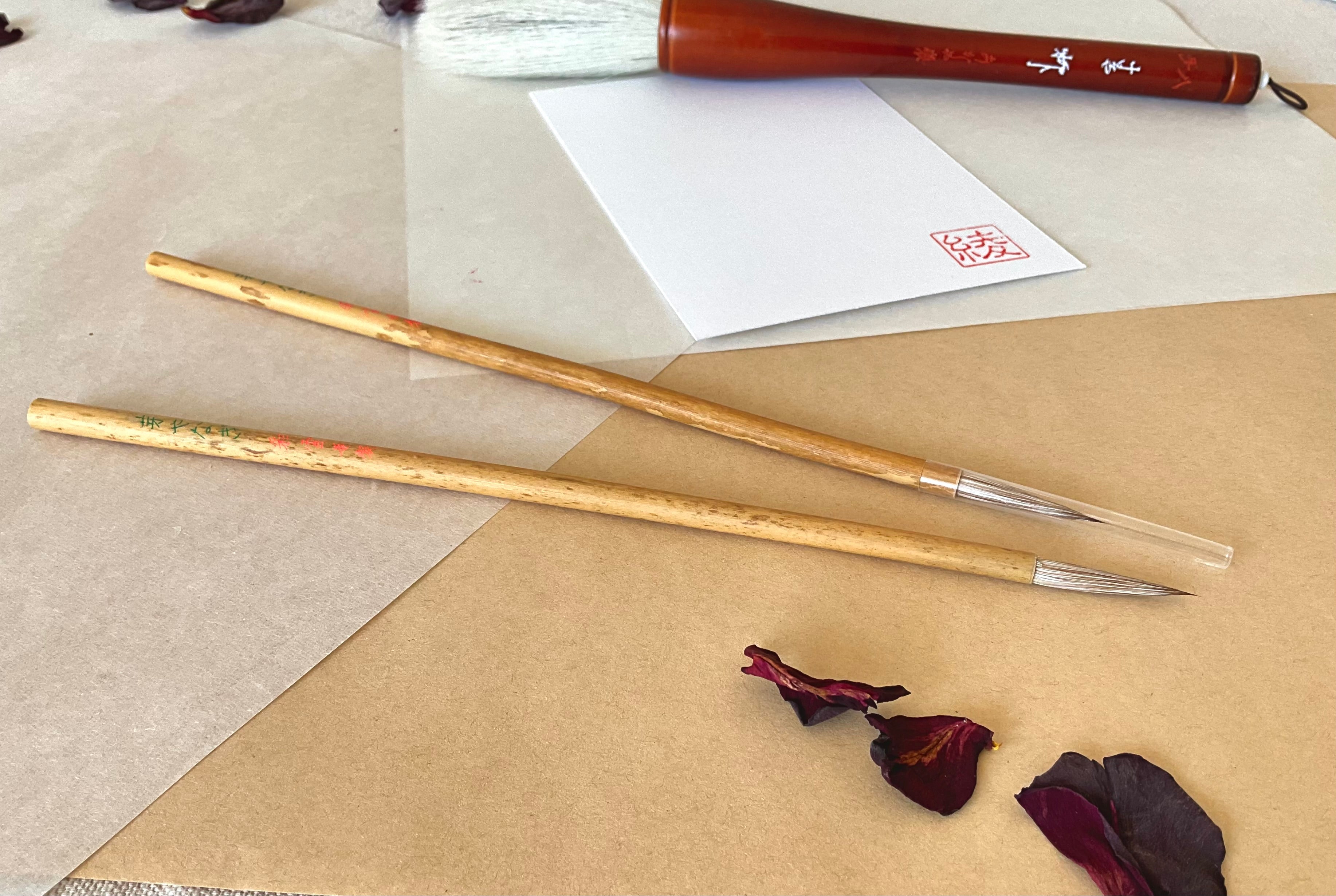 Two Saiundo calligraphy brush with light bamboo handle and fine pointed bristle, one covered with a lid, one is open, resting on layered washi paper with dried rose petals and a red hanko stamp card