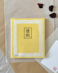 Single pack of Morita Washi kaishi paper at an angle on layered washi paper, yellow washi cover with framed kaishi character stamp, showing thickness of 100 sheets, with dried rose petals in background