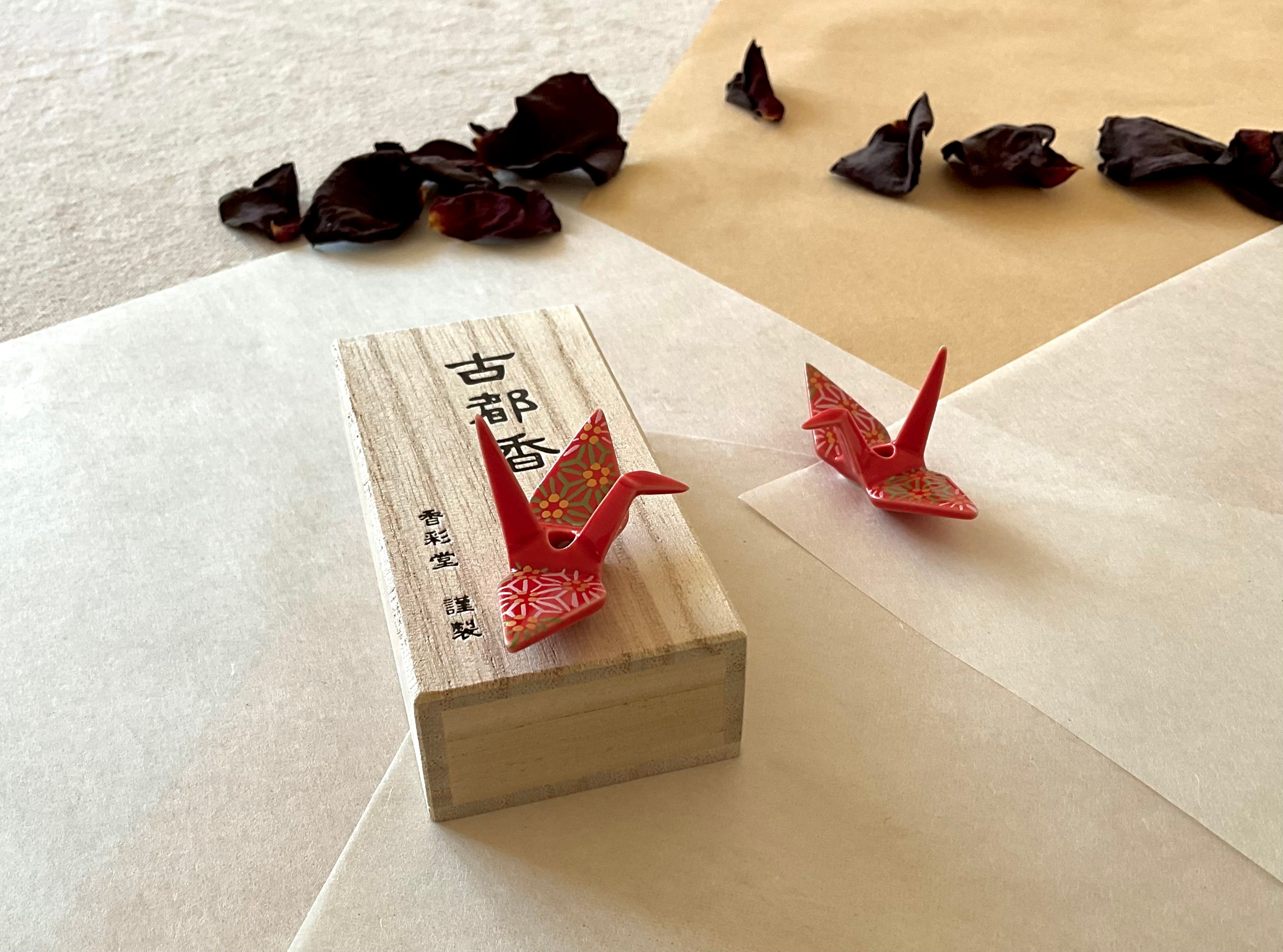 Ceramic origami crane incense holder with red chiyogami pattern glaze standing on a paulownia wood incense box with Japanese kanji, a second crane resting nearby on washi paper, dried rose petals scattered around